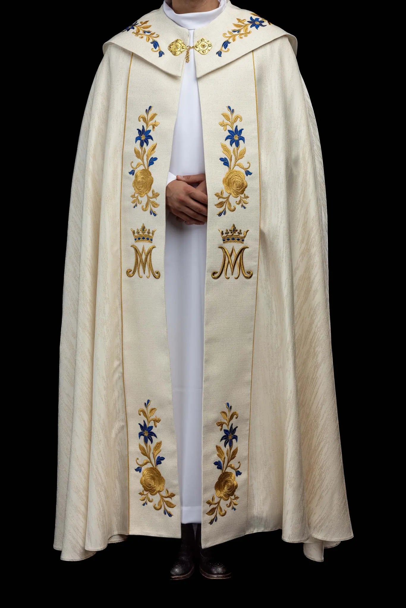 Ecru embroidered cope with Marian symbol, featuring gold and blue floral embroidery, a crowned 'M' monogram, and an ornate gold clasp with chain.