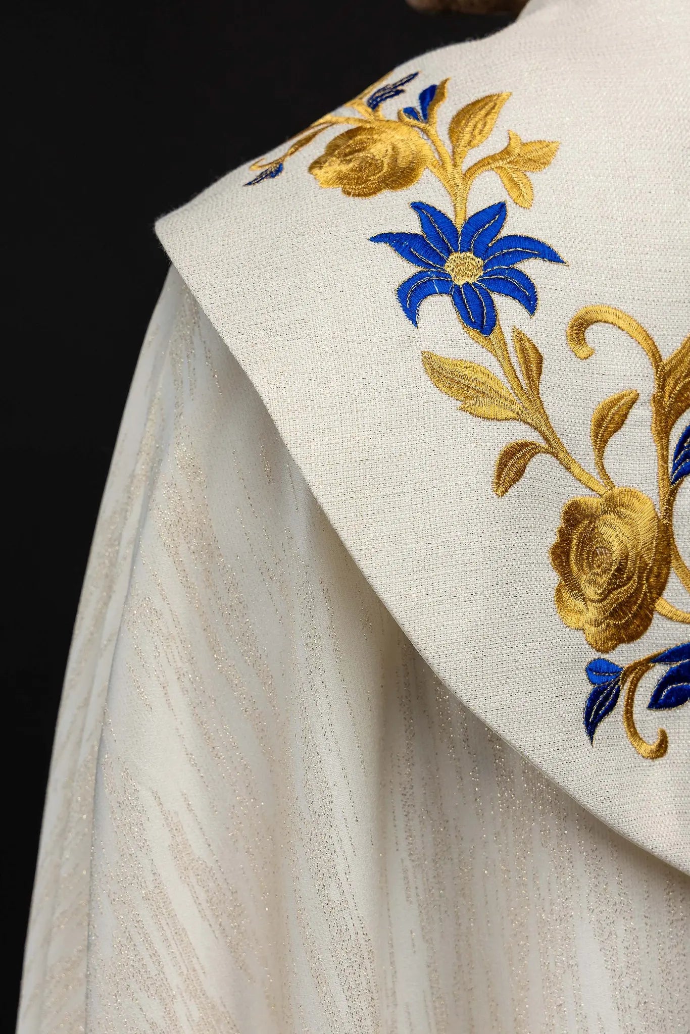 Ecru embroidered cope with Marian symbol, featuring gold and blue floral embroidery, a crowned 'M' monogram, and an ornate gold clasp with chain.